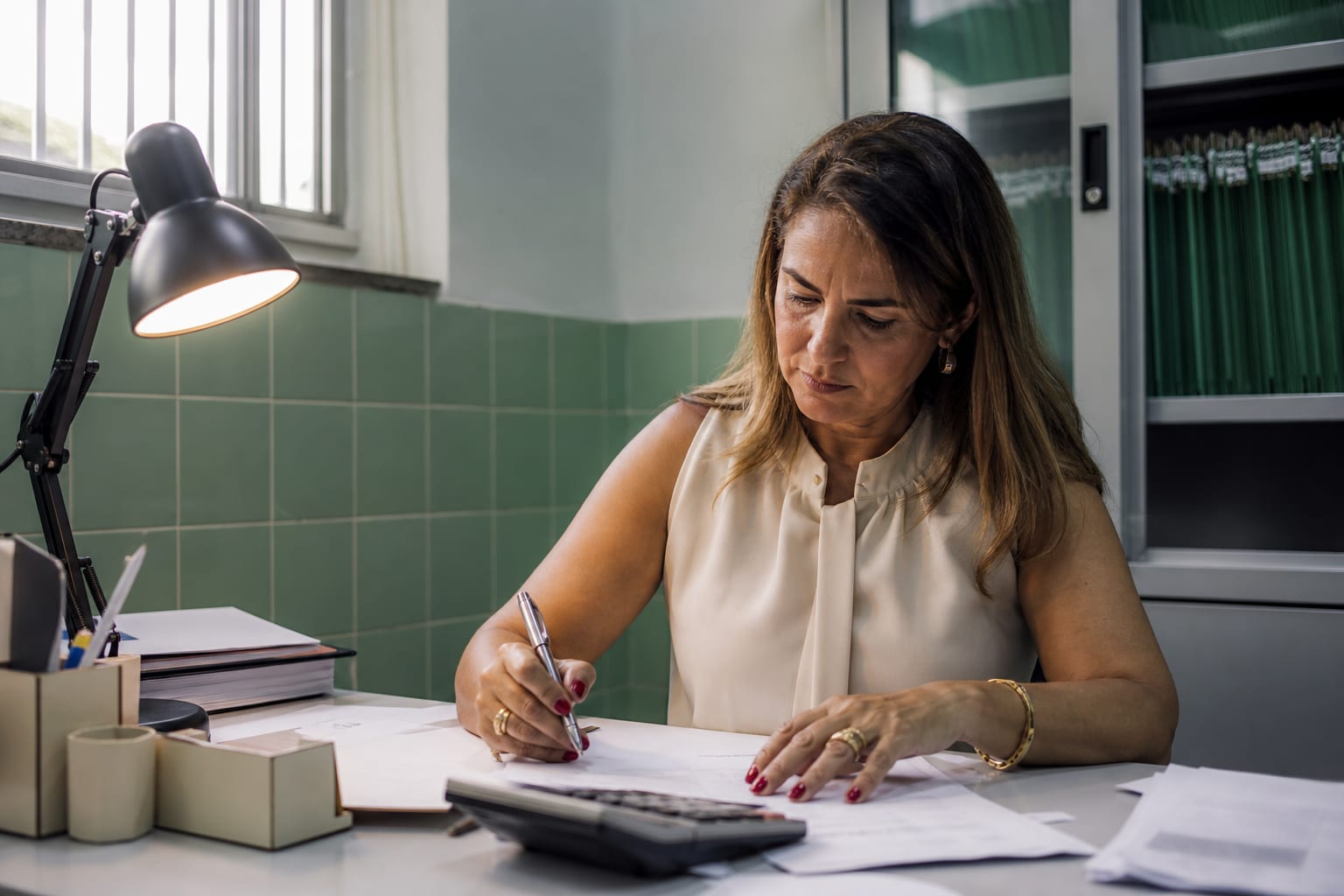 Sócia de clínica faz contas com calculadora de mesa ao lado de papéis impressos na sala de administração